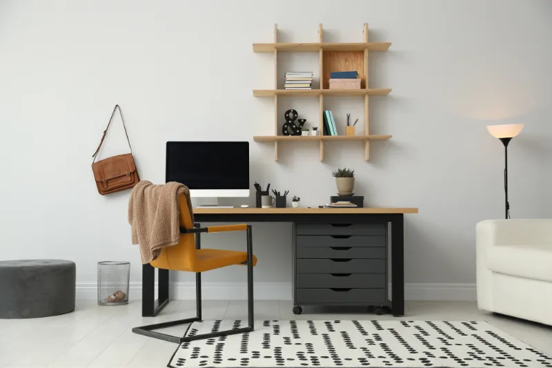 A modern home office with a desk, computer, yellow chair, tan sweater, wall shelves holding books and decor, a brown bag hanging on the wall, a floor lamp, and a white sofa on a patterned rug.